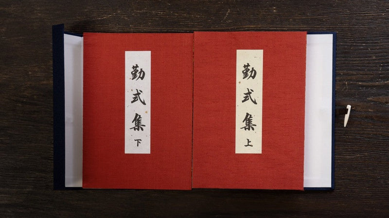 黒い木の台の上で、紺の帙を開いて、赤い表紙の「勤式集」上下2冊を並べたところ